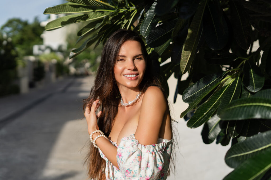Young beautiful woman in a romantic dress with a floral print, and a pearl necklace bracelet, against the backdrop of tropical leaves